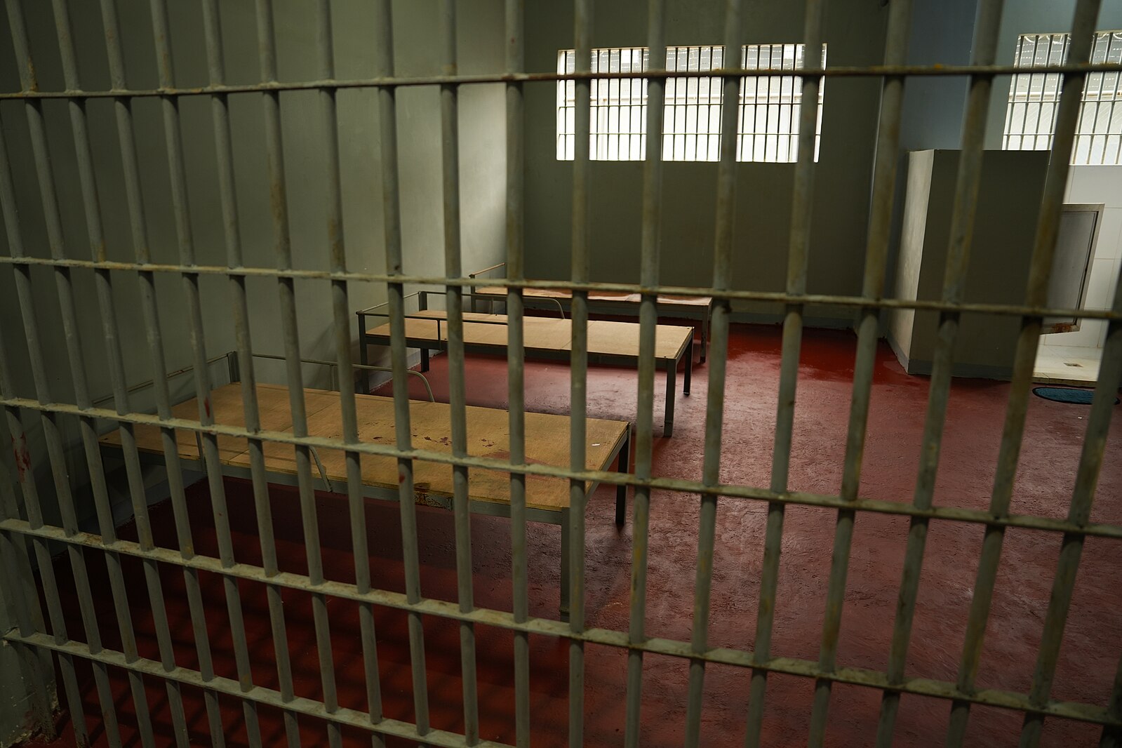 Empty prison cell with barred windows and basic wooden furniture.